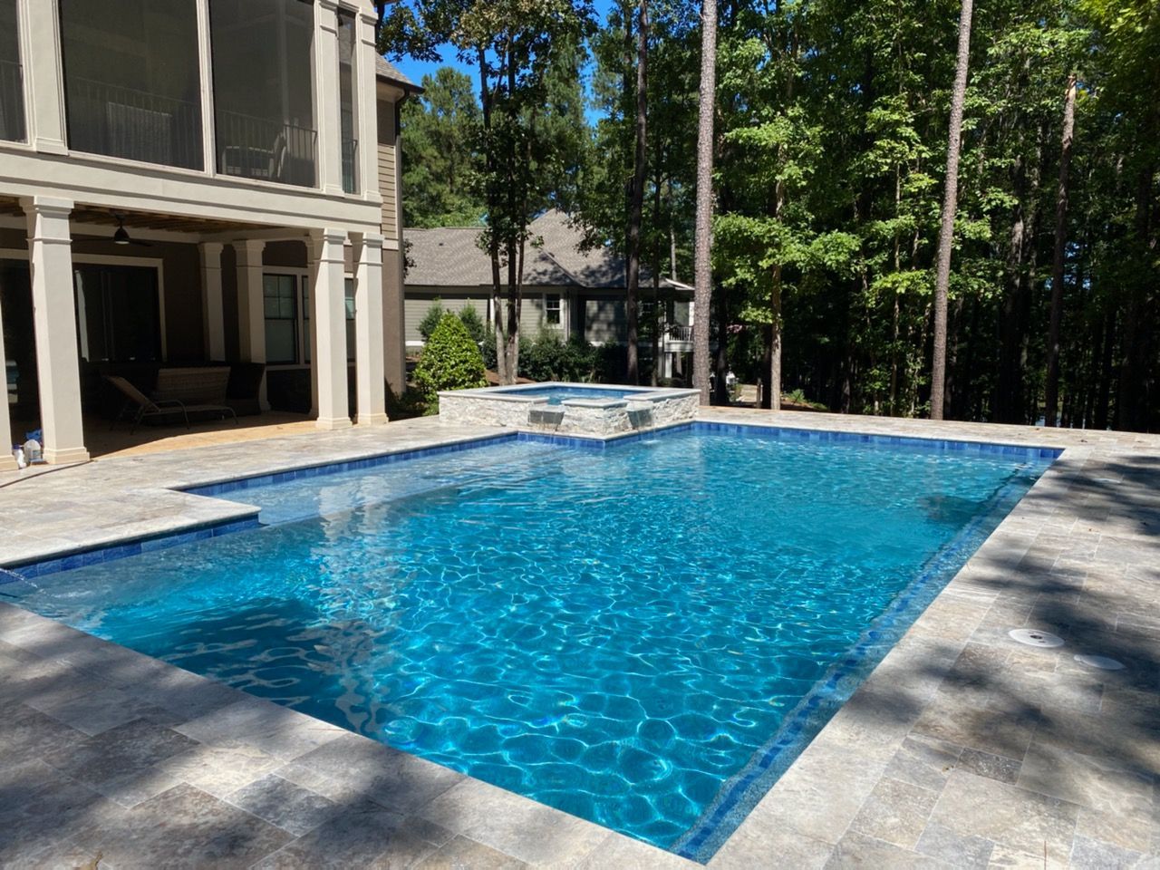Swimming pool with blue tiles surrounded by stone patio next to a house and trees.