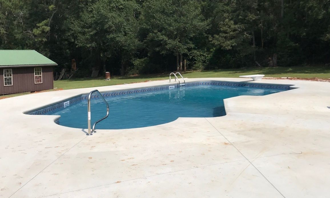 Swimming pool with blue water, surrounded by concrete patio and a small brown shed in a wooded area.