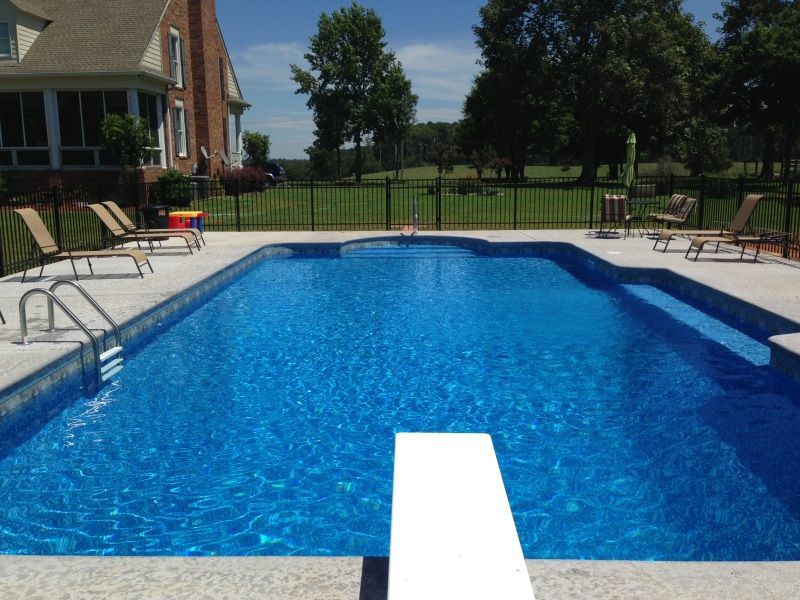Rectangular swimming pool with diving board; house and green lawn in the background on a sunny day.