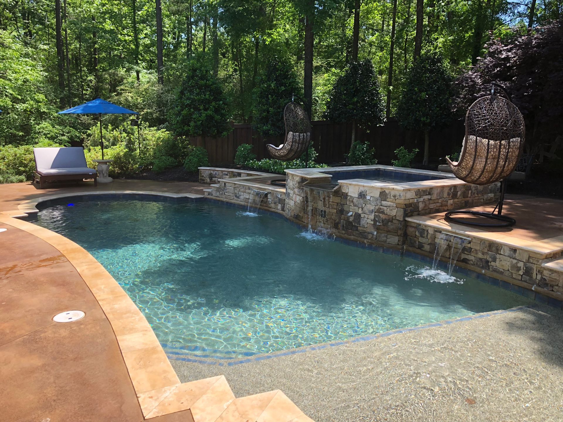Pool with waterfall, spa, and seating area surrounded by trees.