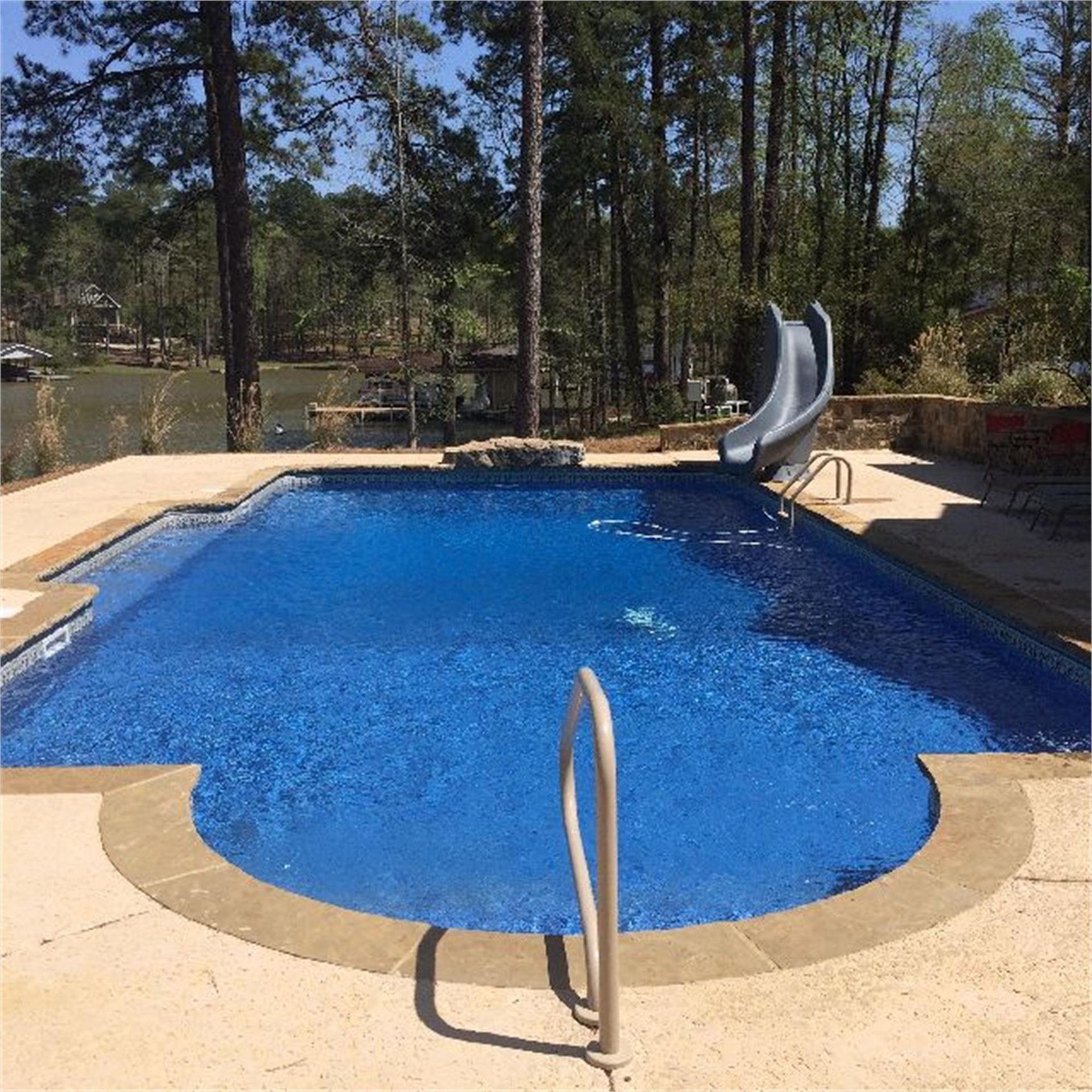 Swimming pool with blue water, a water slide, and a view of trees and a lake.
