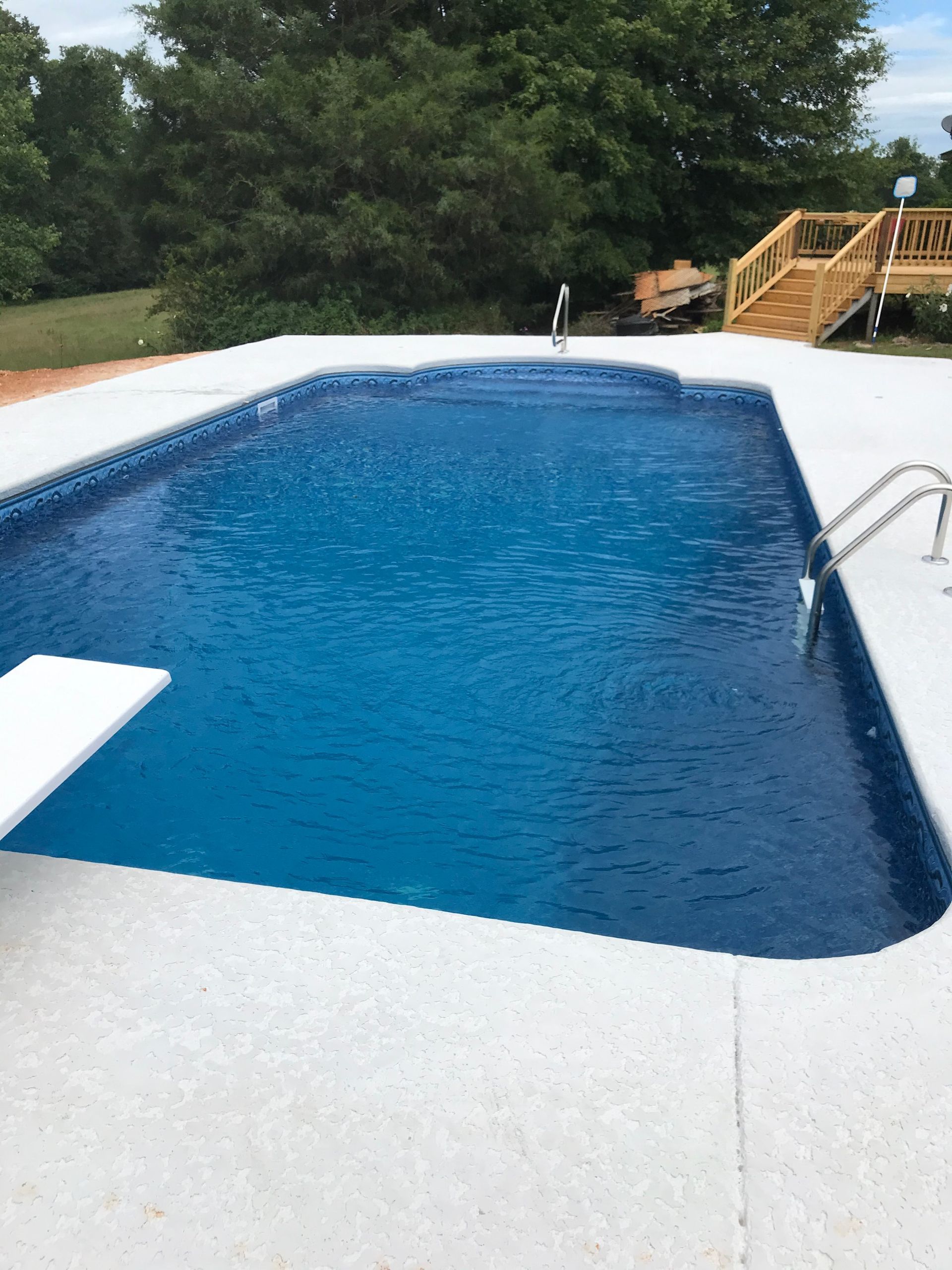 Blue swimming pool with white concrete surround and diving board. Wooden deck in background.