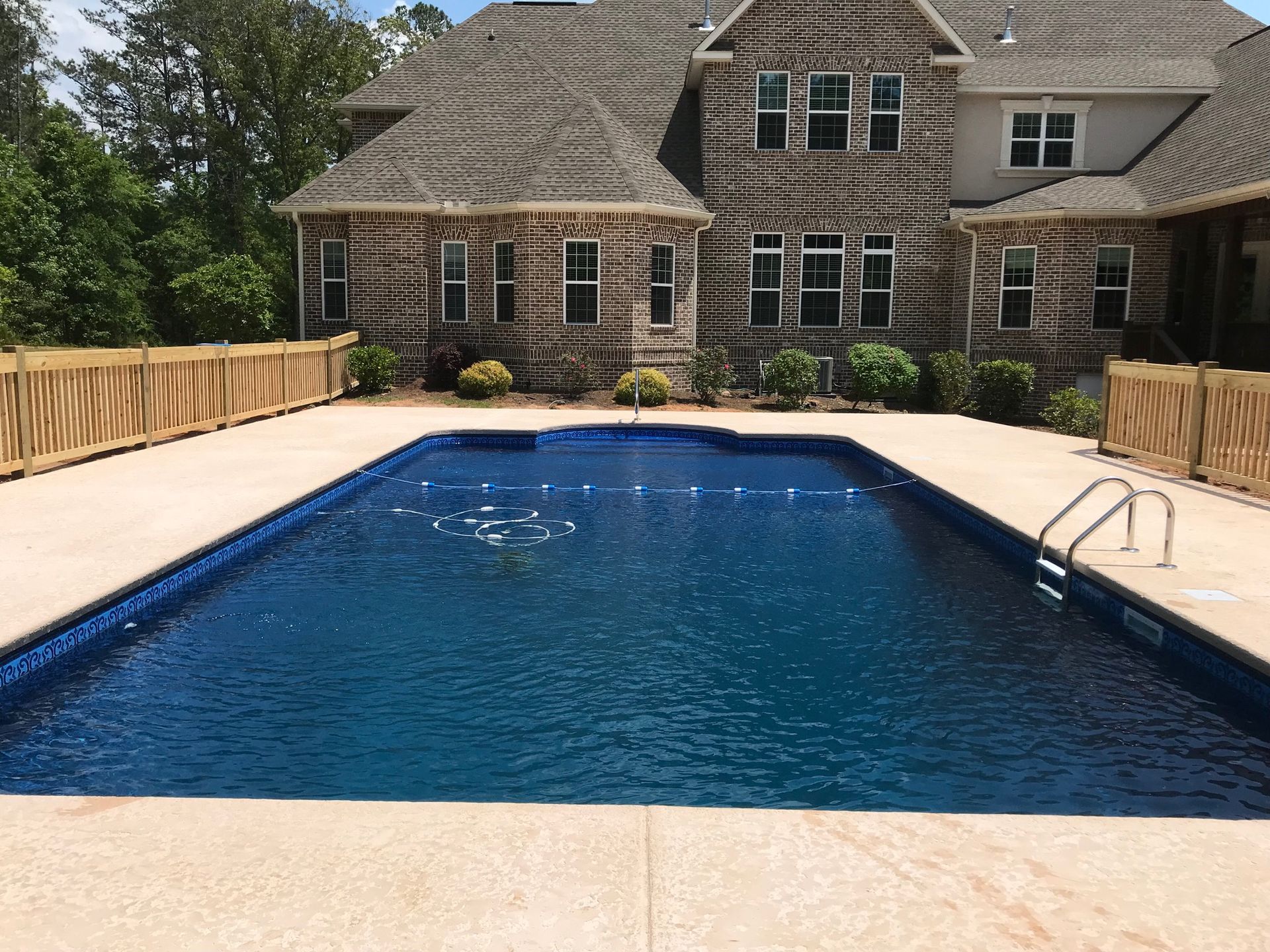 Rectangular pool with blue water in front of a brick house, surrounded by a beige patio and wooden fence.