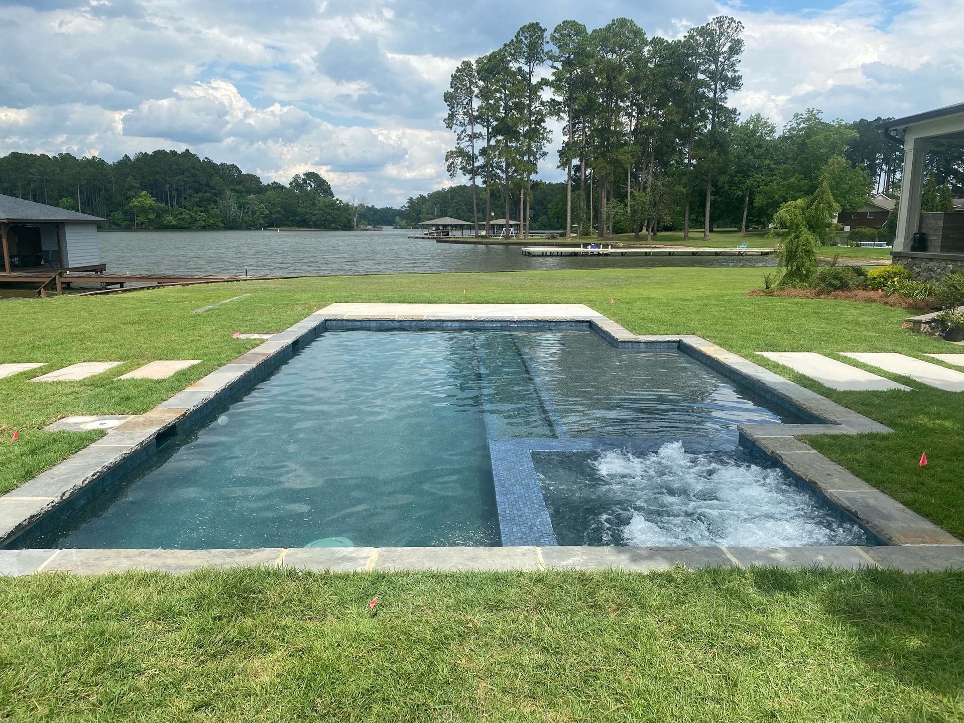 A rectangular pool with a spa, surrounded by grass and a lake backdrop under a cloudy sky.
