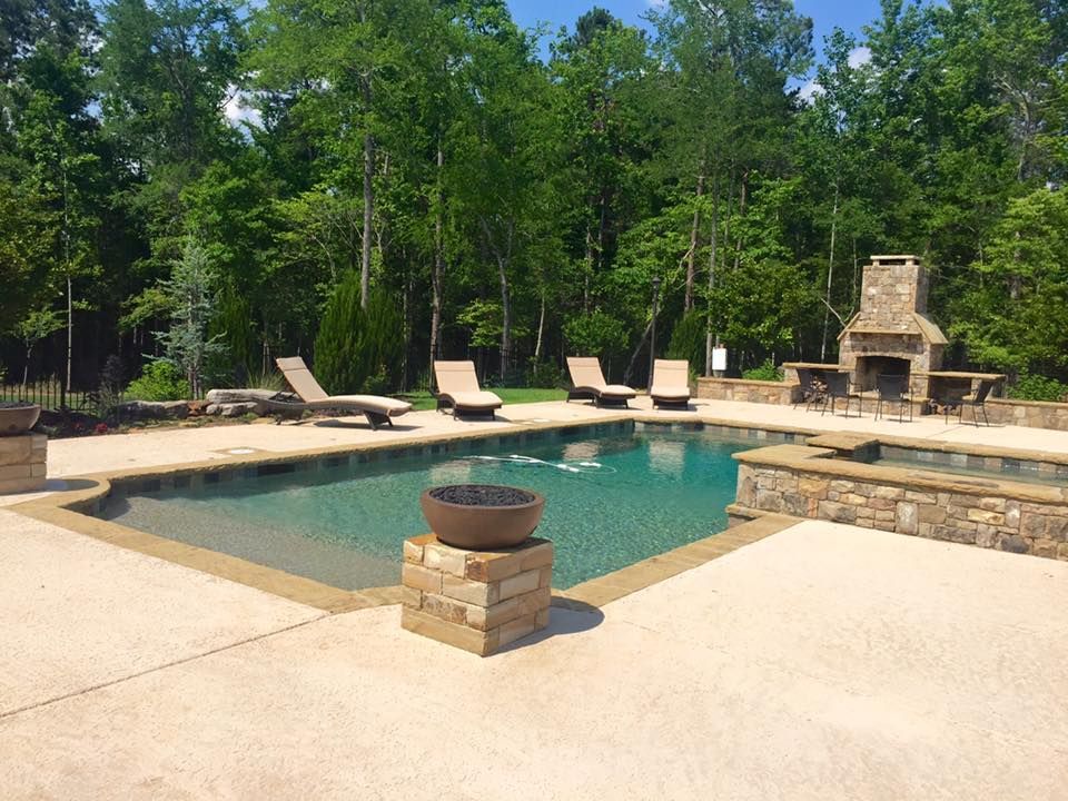 Pool area with lounge chairs, stone fireplace, and a bowl on a pedestal. Green trees in the background.
