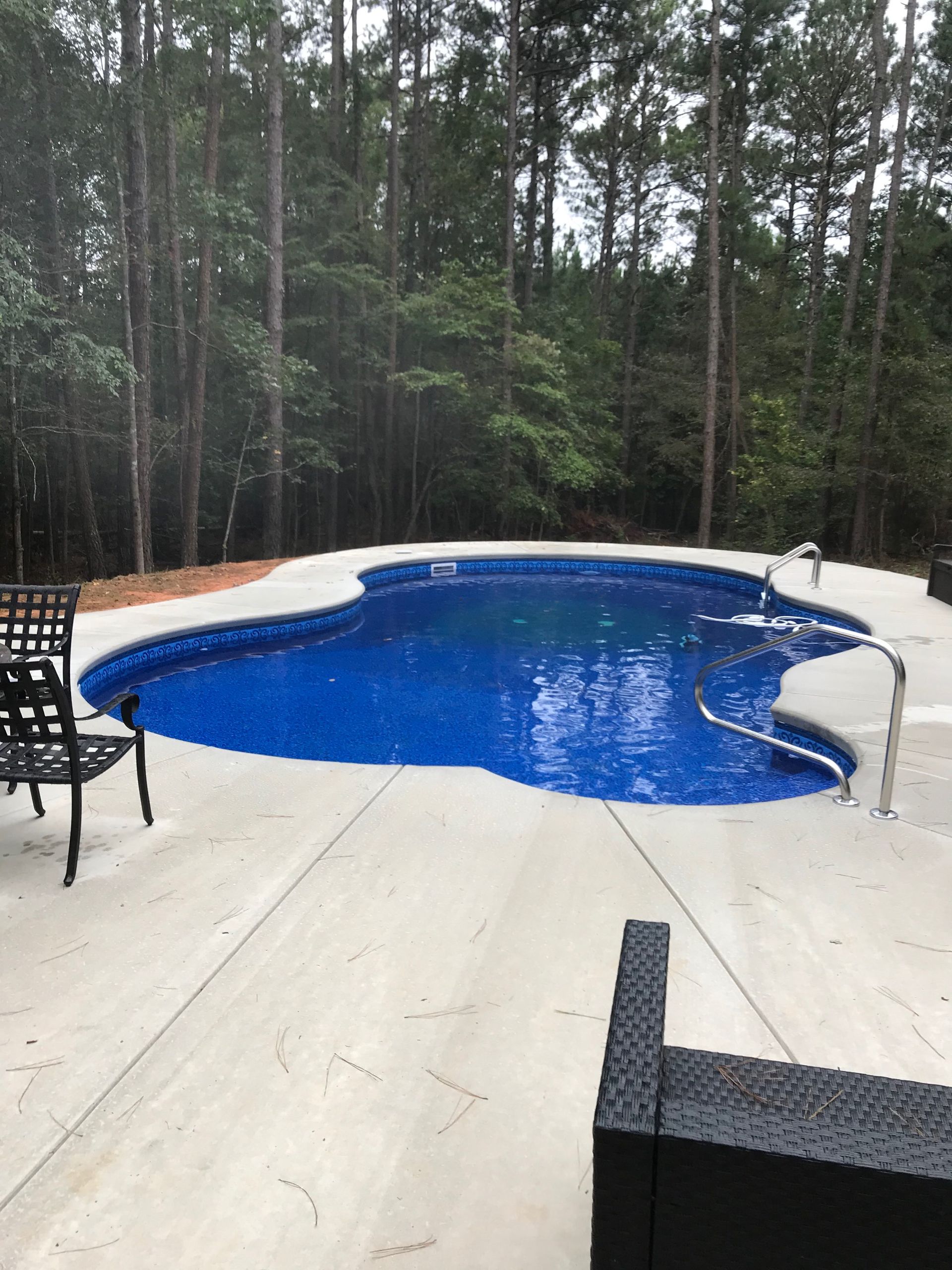 Blue swimming pool surrounded by a concrete patio, with forest in the background.
