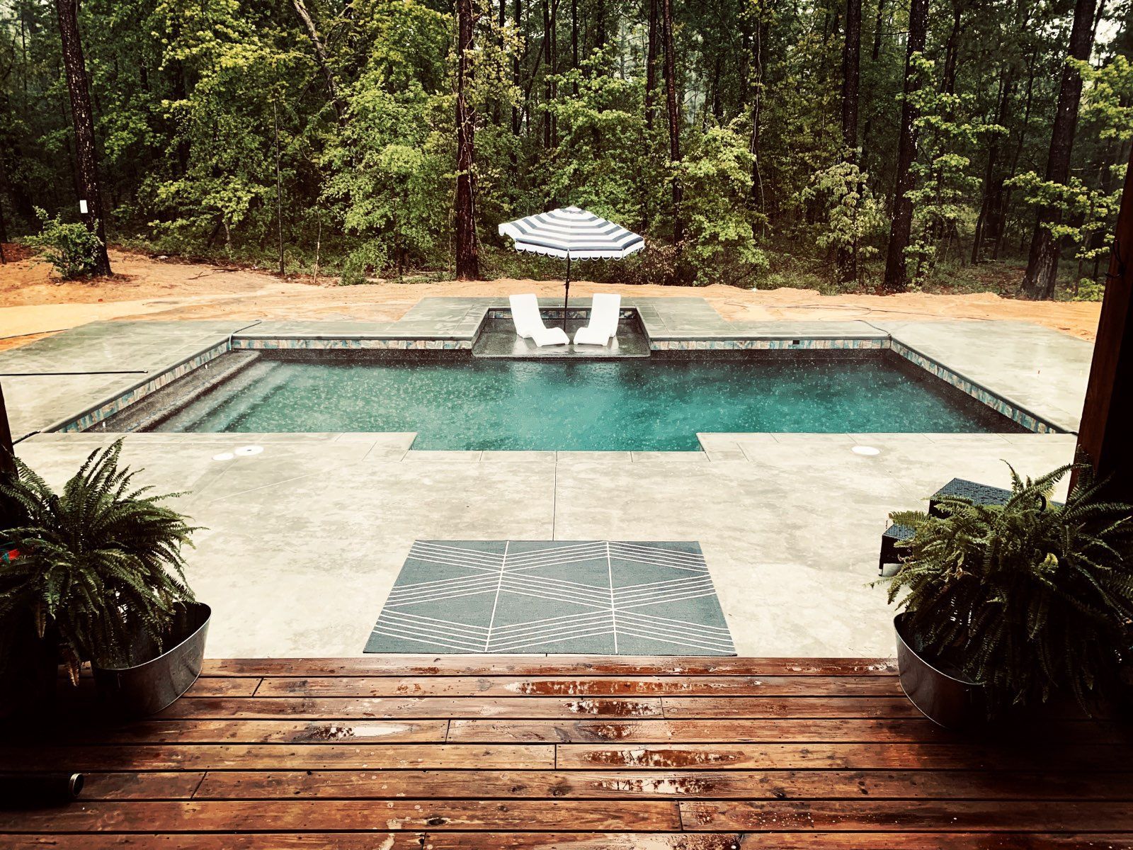 Outdoor pool surrounded by trees. Two white chairs with striped umbrella. Ferns in planters.