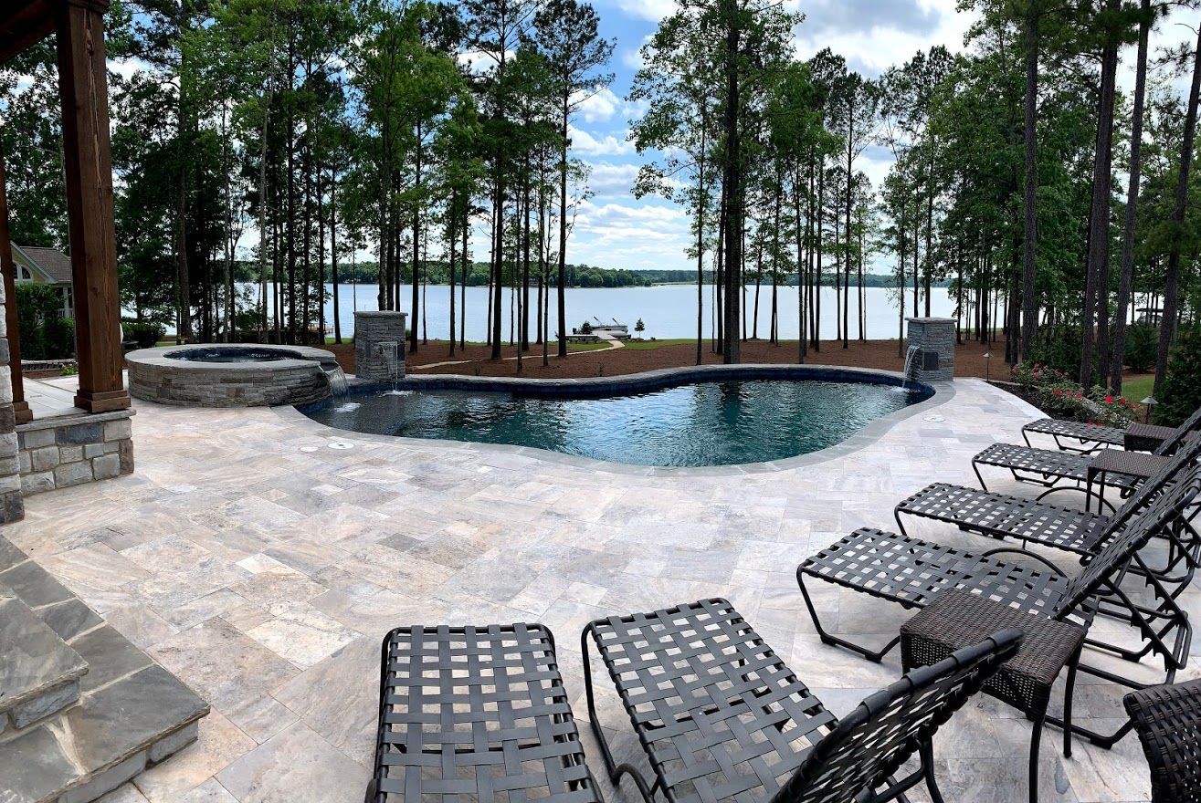 Poolside patio with lounge chairs overlooking a lake and trees under a cloudy sky.