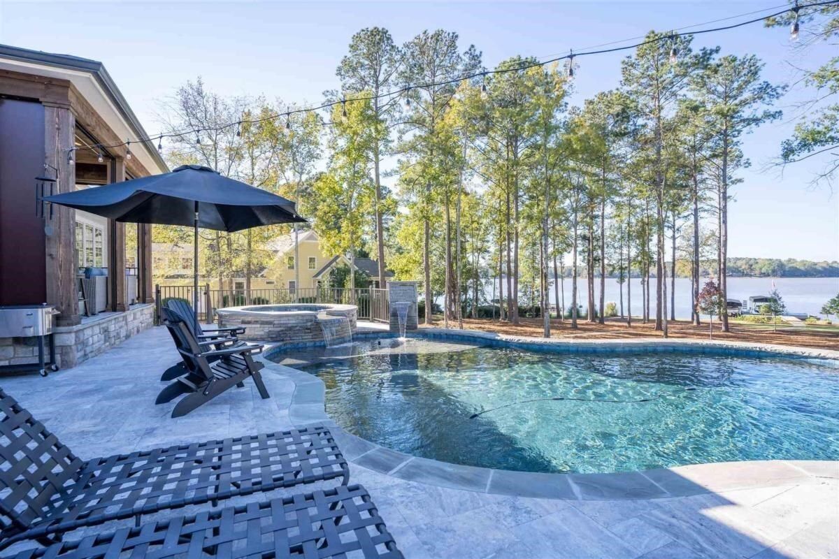 Poolside view with lounge chairs, a black umbrella, and a clear pool overlooking a lake.
