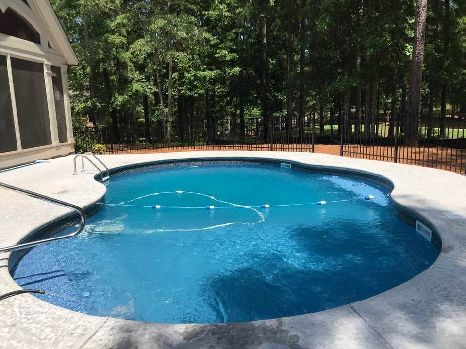 Swimming pool with blue water, surrounded by a concrete deck, next to a house and trees.