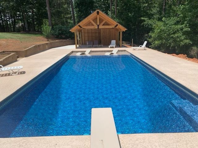 Rectangular pool with diving board, cabana, blue water, and concrete patio, set in a wooded backyard.