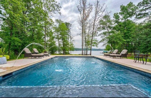 Swimming pool with fountains, lounge chairs, and view of a lake surrounded by trees.