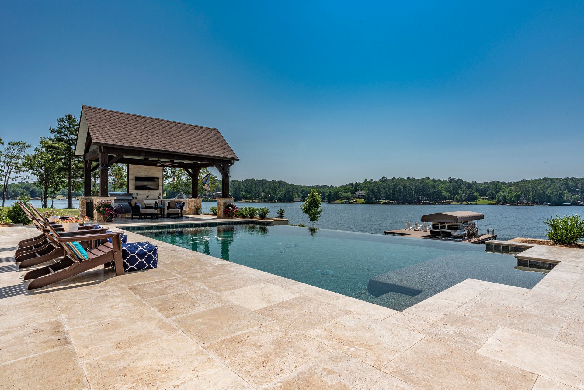 Infinity pool next to a lake with a covered seating area on a sunny day.