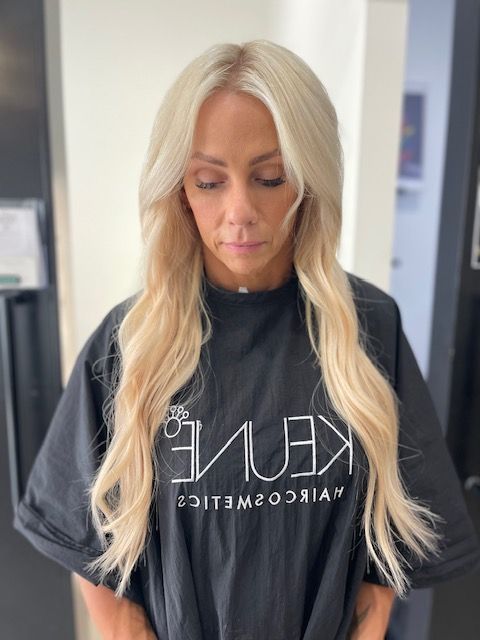 Woman with long blonde hair wearing a black salon cape, looking down in a bright salon setting