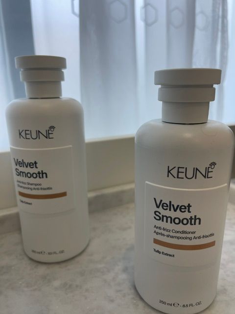 Two KEUNE Velvet Smooth hair product bottles on a countertop in front of sheer curtains.