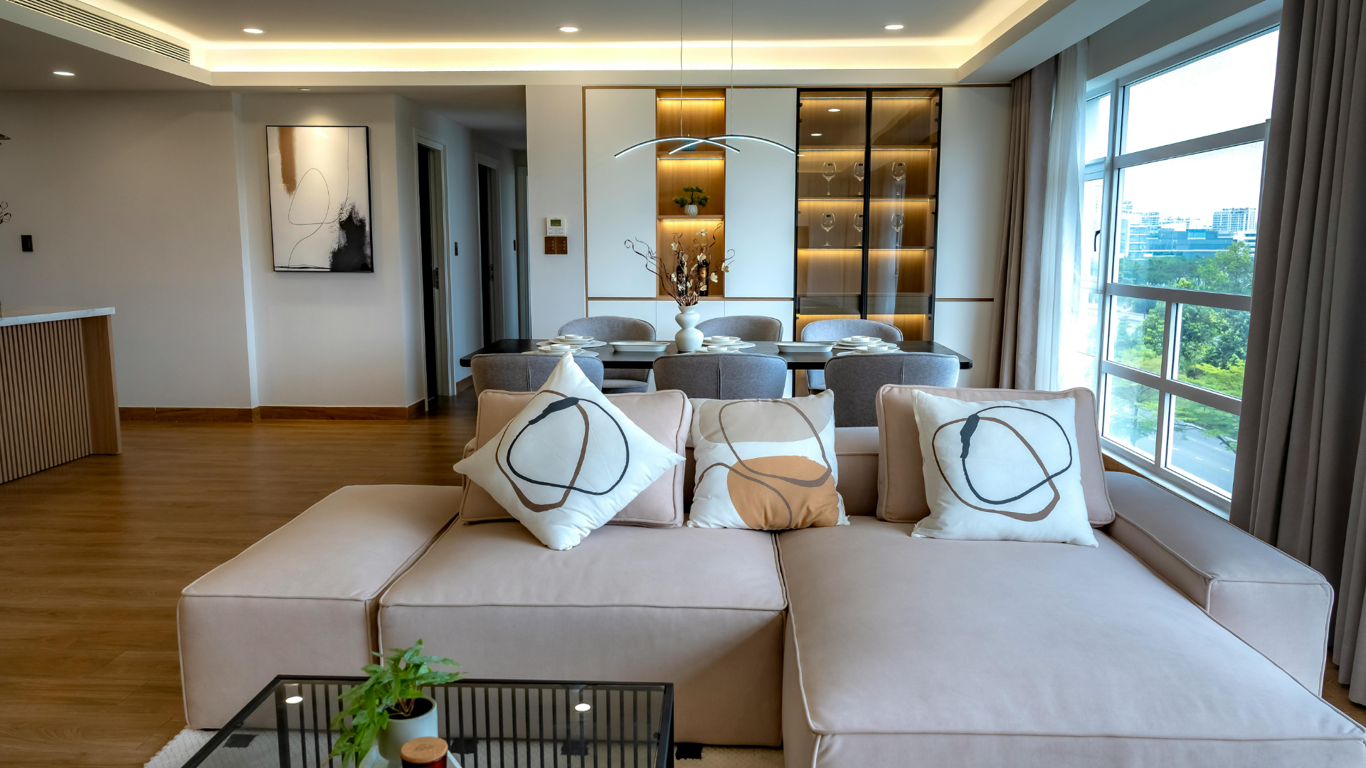 Modern living room with beige sectional sofa, wood floors, and large windows overlooking greenery