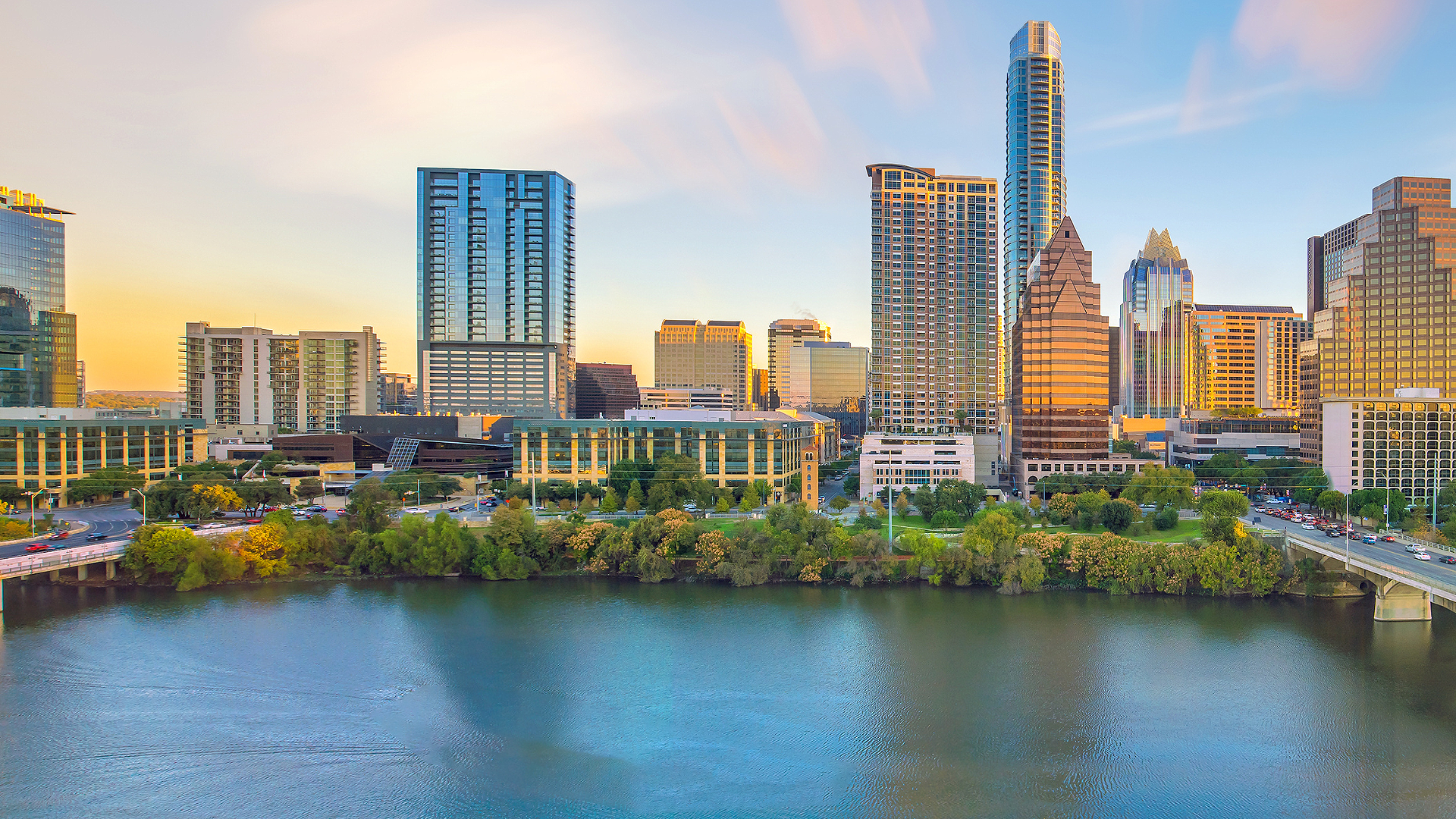 City skyline at sunset reflected over a calm river, with modern high-rises and a bridge in view