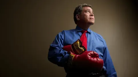A man in a blue shirt and red tie is holding a pair of red boxing gloves.