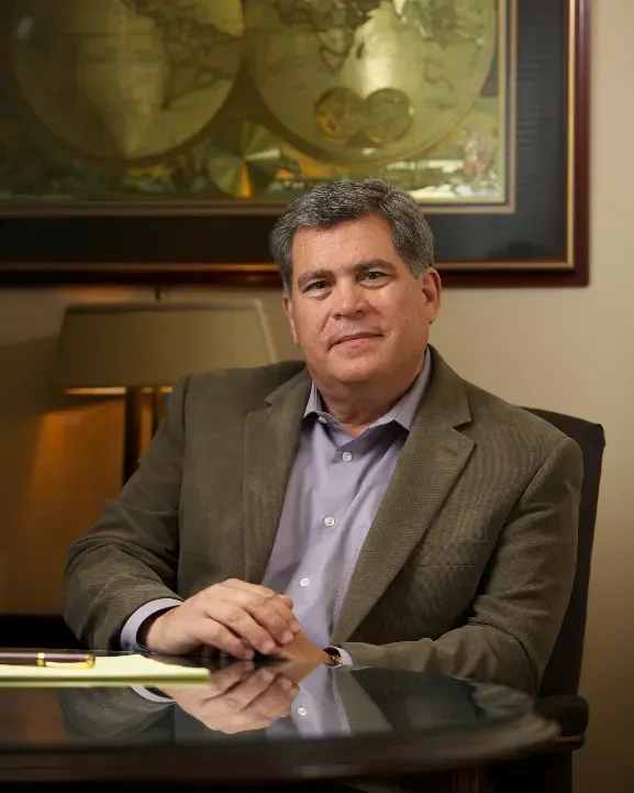 Attorney Ralph G Martinez in a suit sits at a desk in front of a map of the world