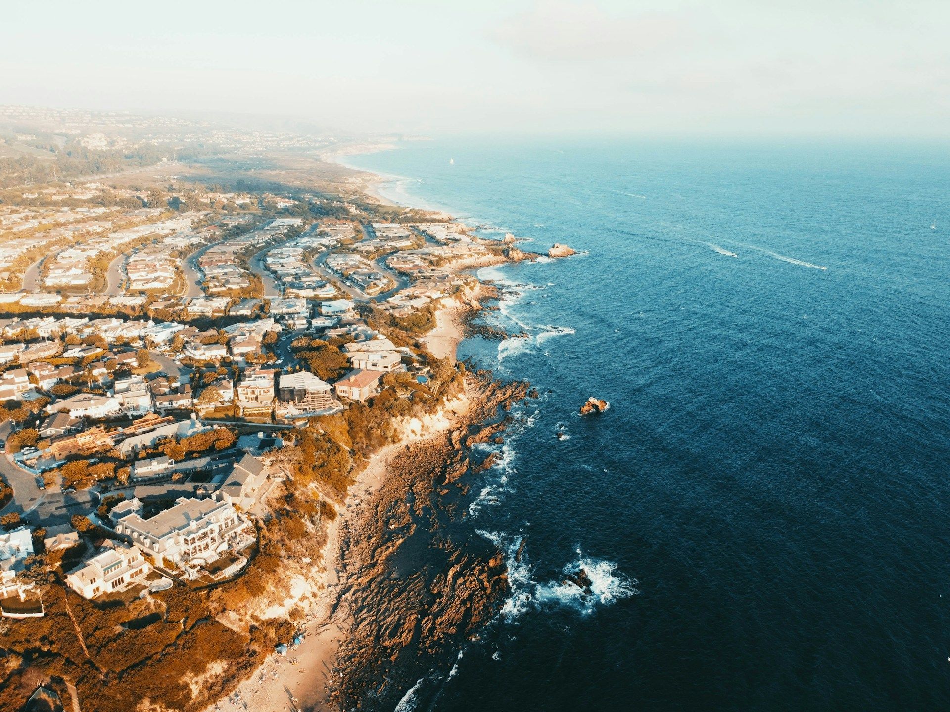 Coastal aerial view: Houses and beach meet the blue ocean under a hazy sky.
