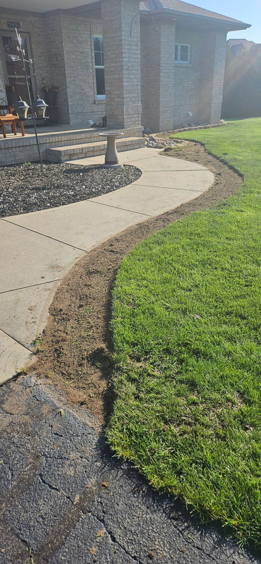 Cracked driveway and uneven concrete sidewalk leading to a brick house with a green lawn.