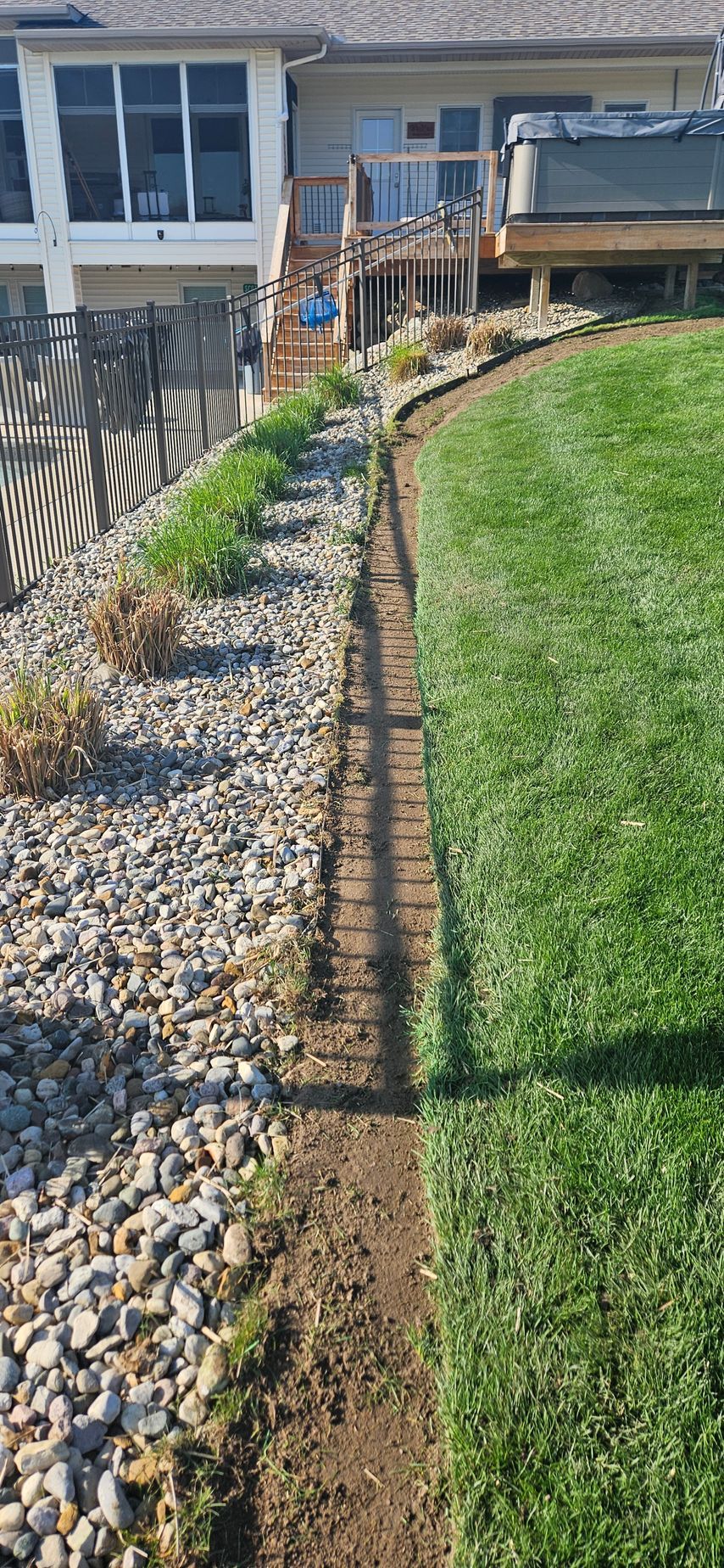 Curved landscaped rock bed beside a green lawn and house patio with wooden railing