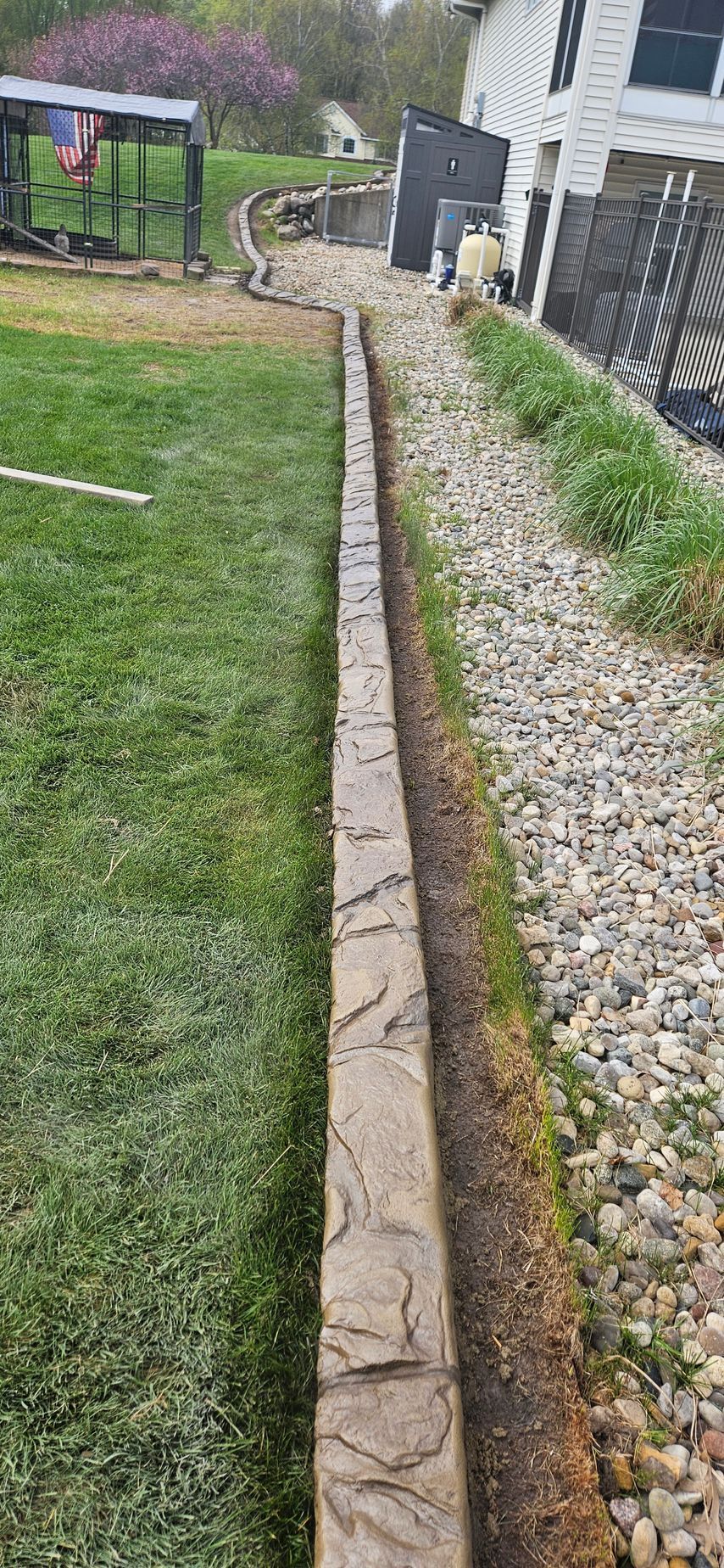 Narrow stone walkway beside a grassy yard and gravel border leading to a house and fence.
