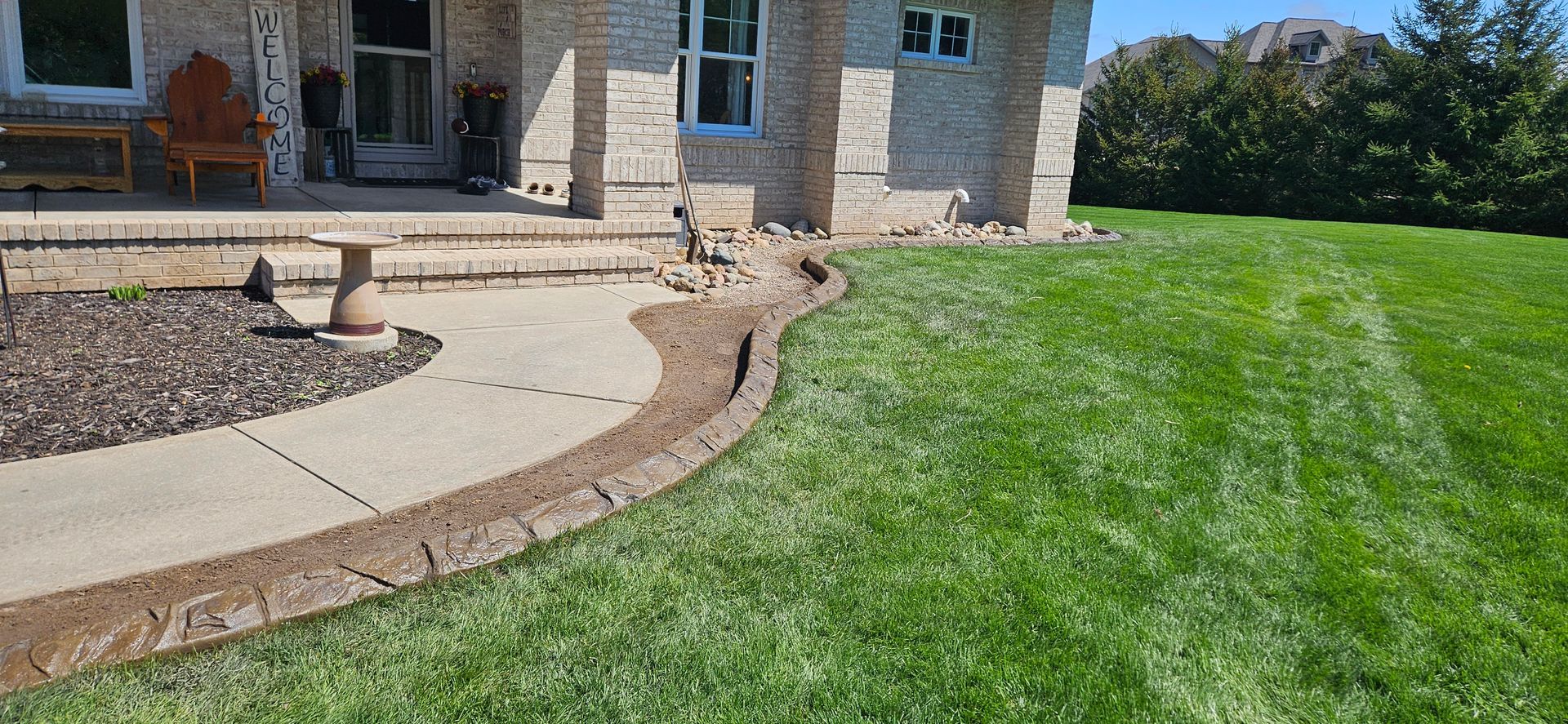 Front yard with curved concrete walkway, stone house porch, and green lawn on a sunny day