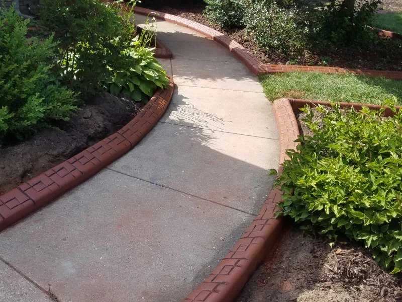 A concrete garden path edged with decorative red brick borders, flanked by green bushes and mulch.
