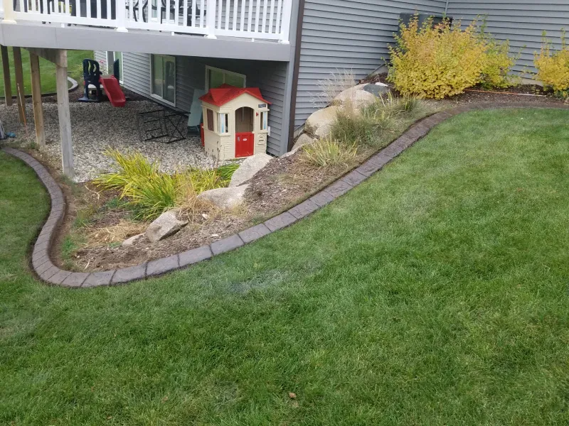 A brown concrete landscape border separates a grassy lawn from a mulch bed with rocks, shrubs, and a playhouse.