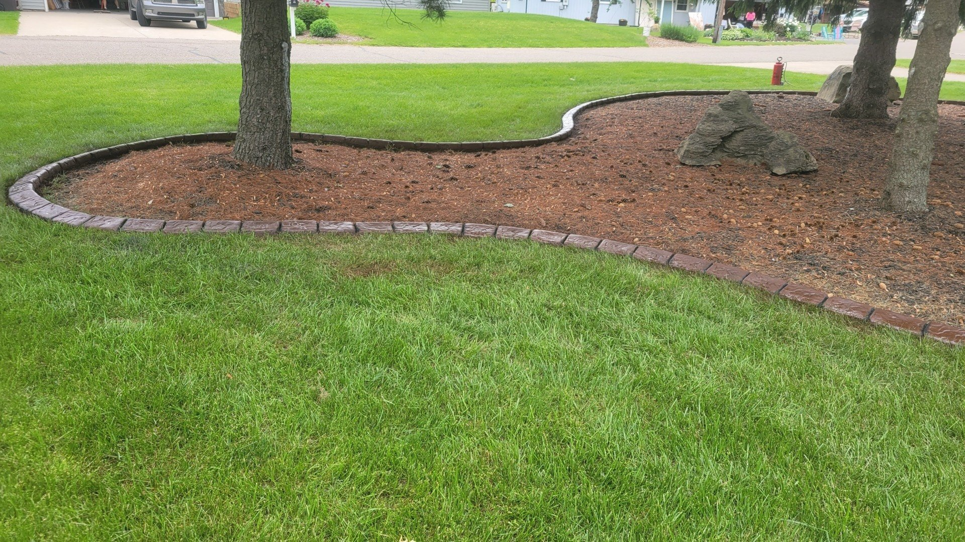 A mulch garden bed with a stone border curves through a green lawn, featuring two tree trunks and a large landscape rock.