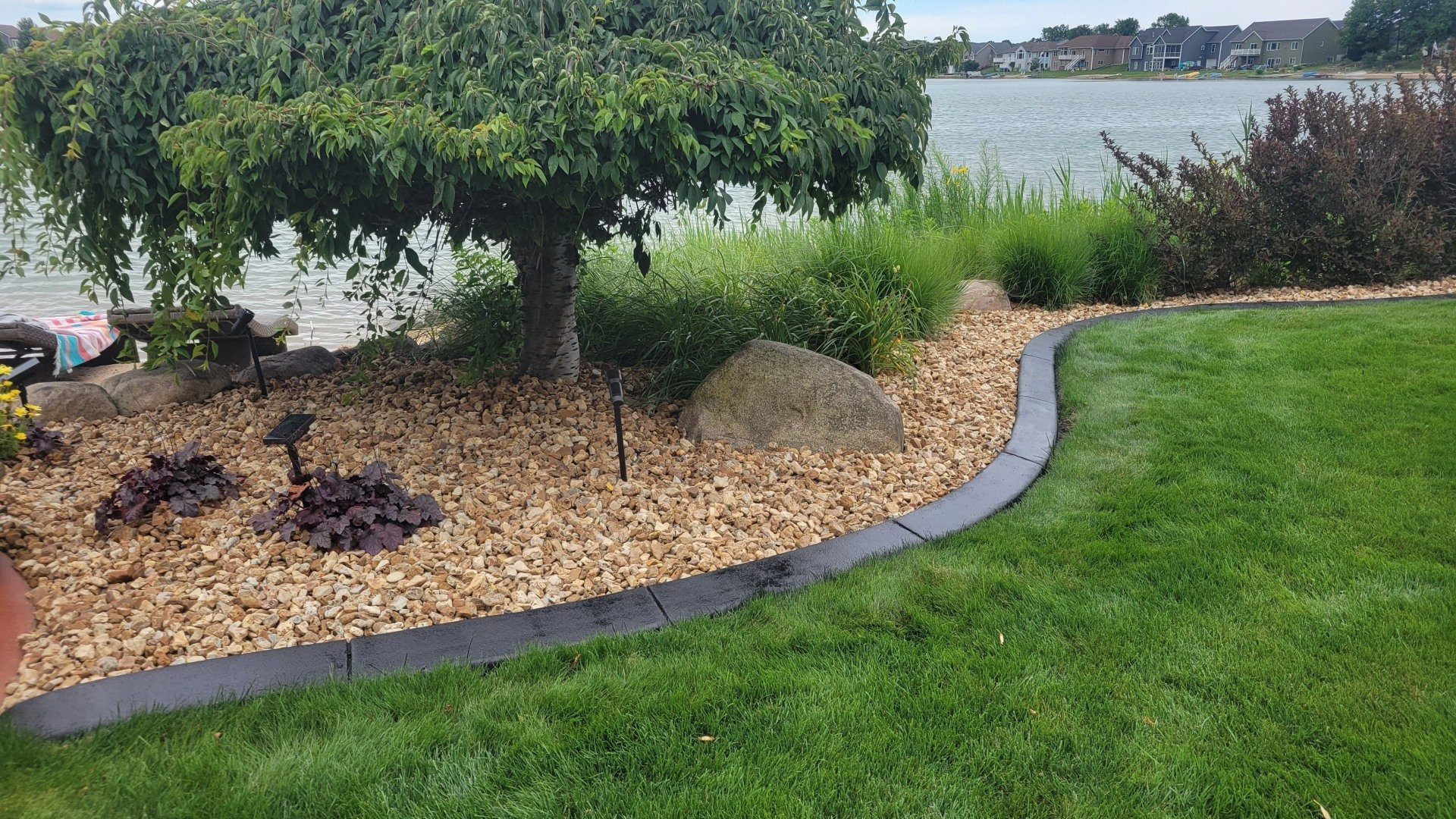 A landscaped garden bed featuring a small tree, river rocks, and a dark stone border, overlooking a calm lake.