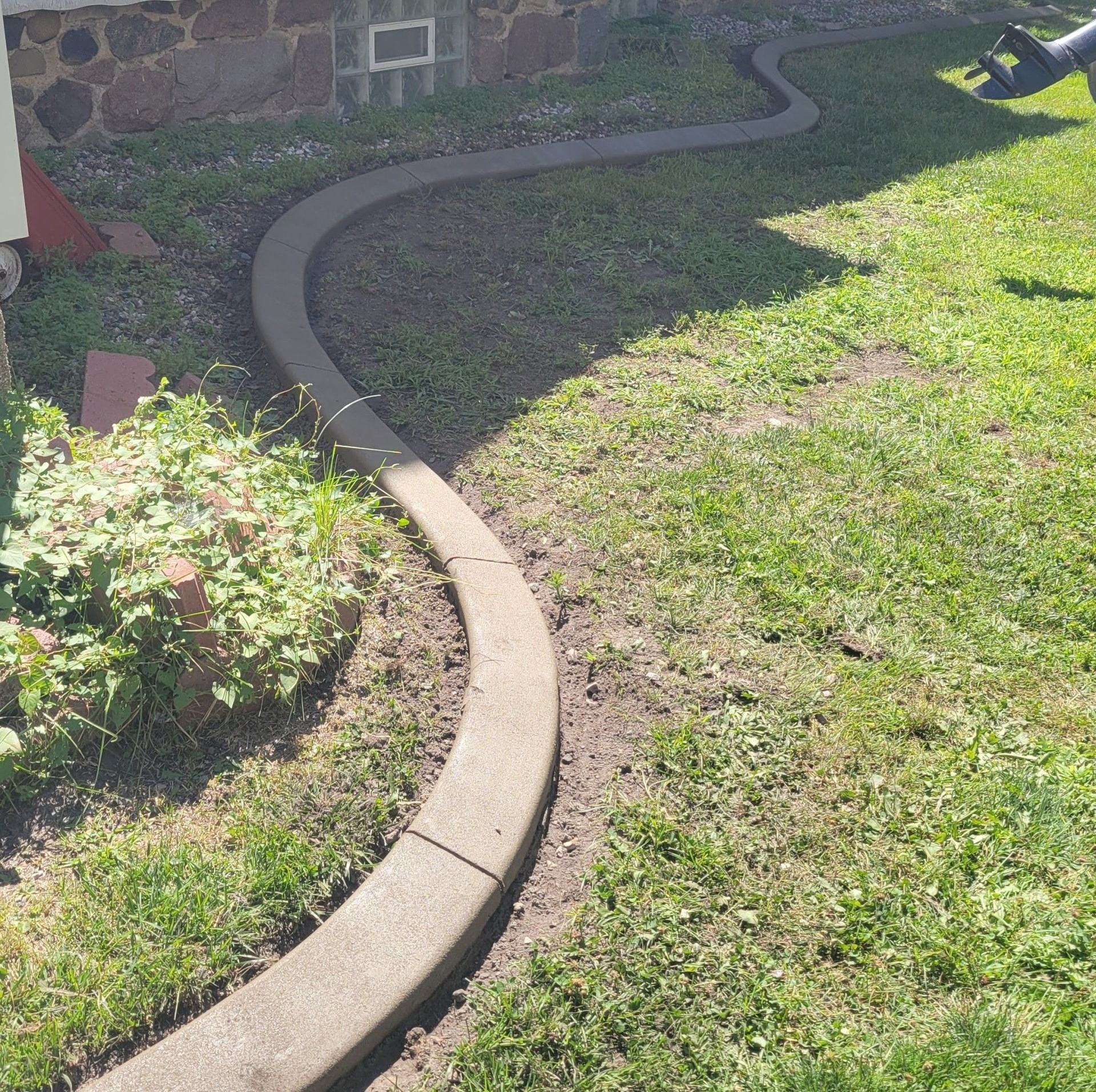 A concrete landscape border edges a garden bed next to a stone house wall and a grassy lawn.