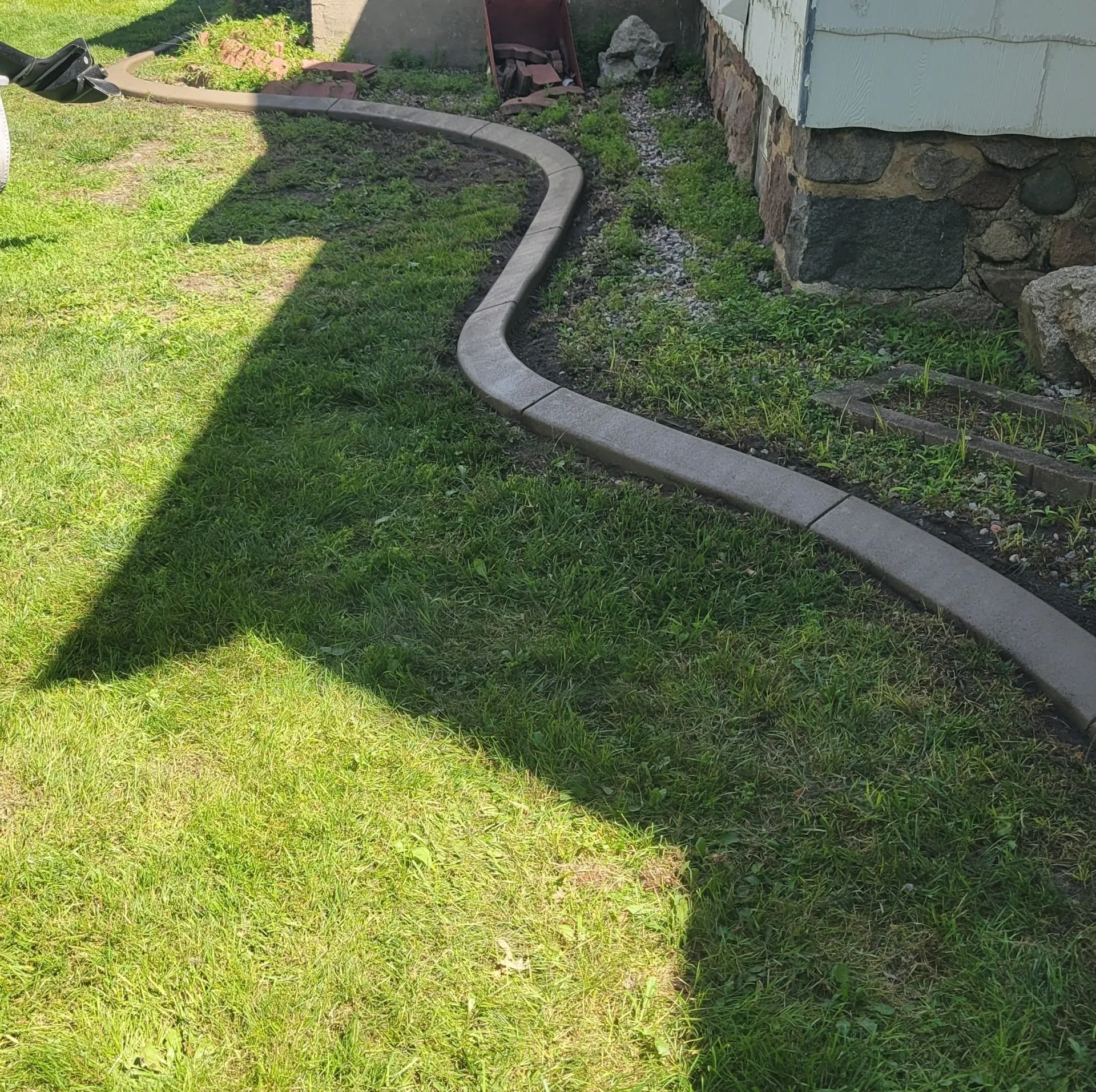 A curved concrete border separates a green grass lawn from a house foundation and patch of dirt.