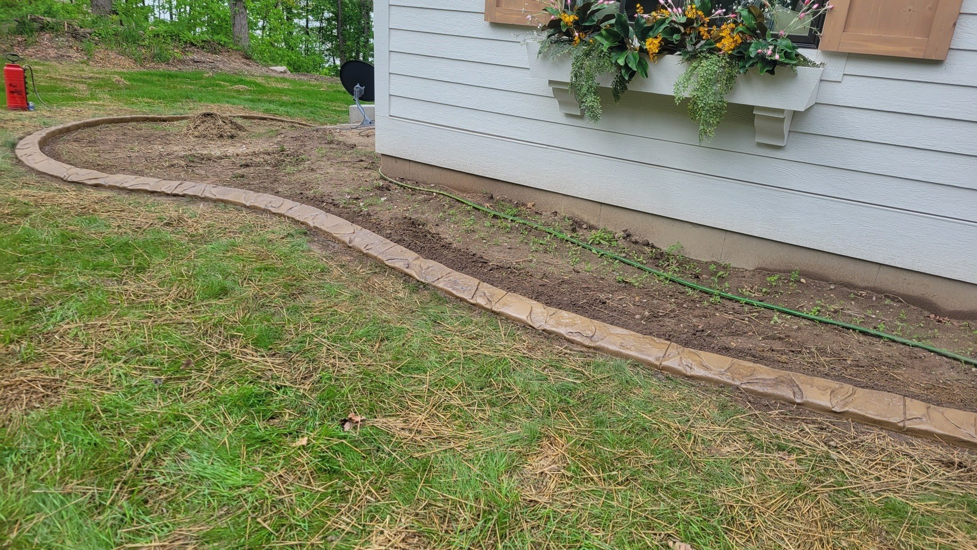 A curved stone garden border separates a patch of bare soil from a lawn, next to a white house with a window box.