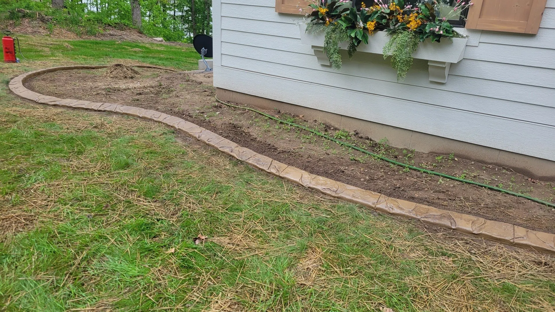 A white house exterior with a flower box under a window, featuring a curved paver border around a soil-filled garden bed.