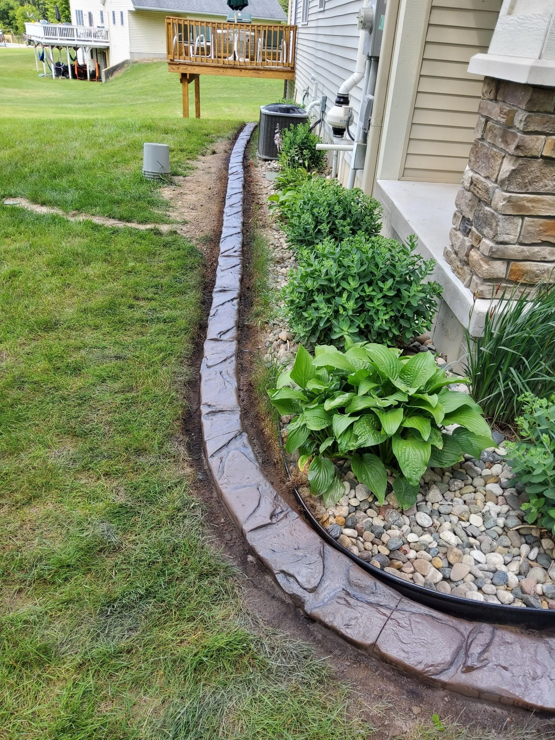 A stamped concrete garden border curves along the side of a house, separating a grassy lawn from a gravel-filled bed.