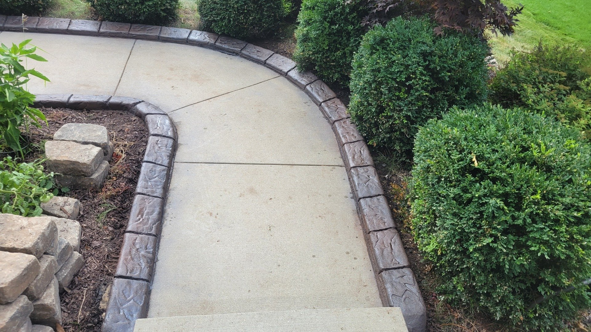 A curved concrete walkway bordered by decorative stone edging, flanked by green bushes and landscape mulch.
