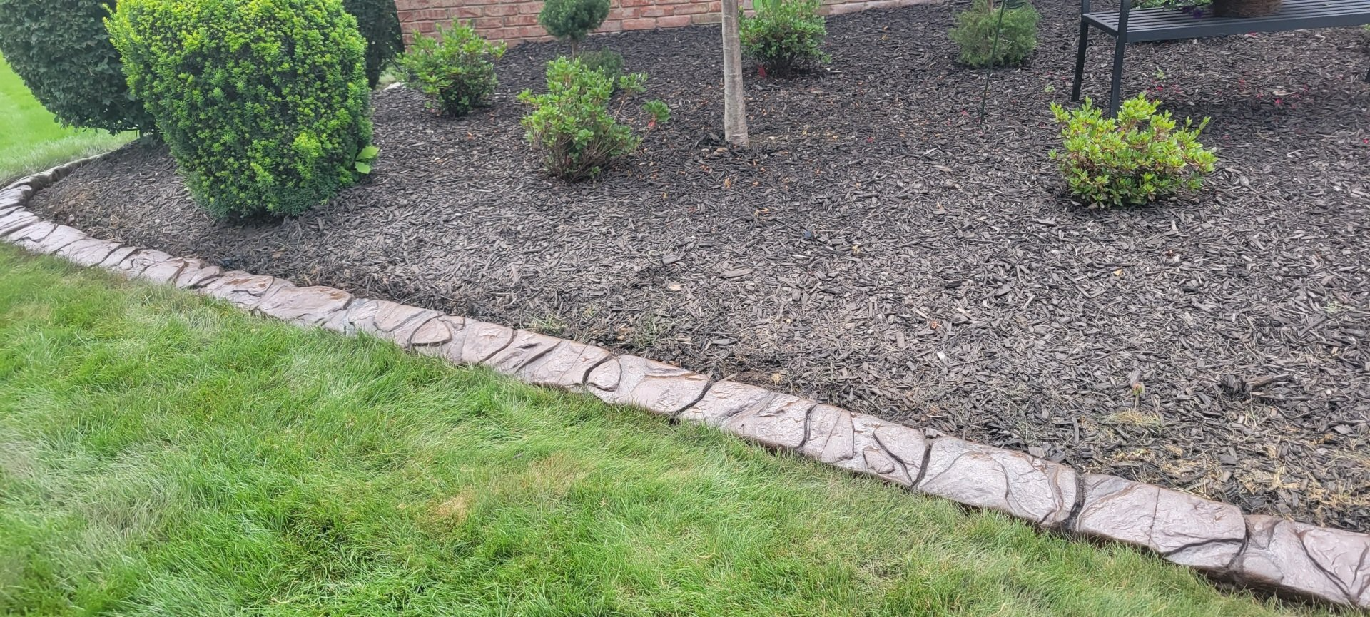 A low-angle view of a decorative stamped concrete garden border separating green lawn from dark wood-mulched flower beds.