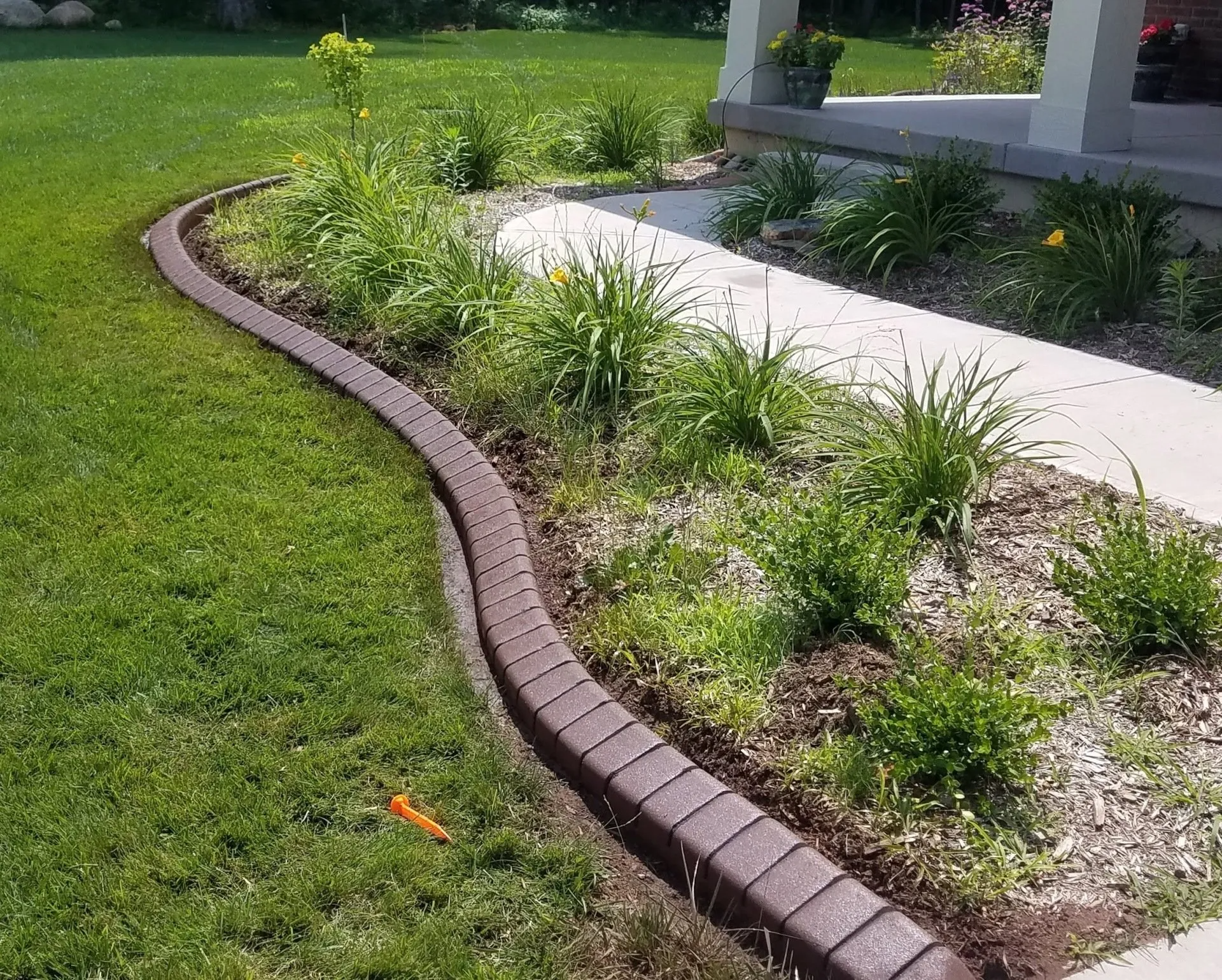 A curved brick garden border separates a green lawn from a mulched flower bed and a concrete walkway.