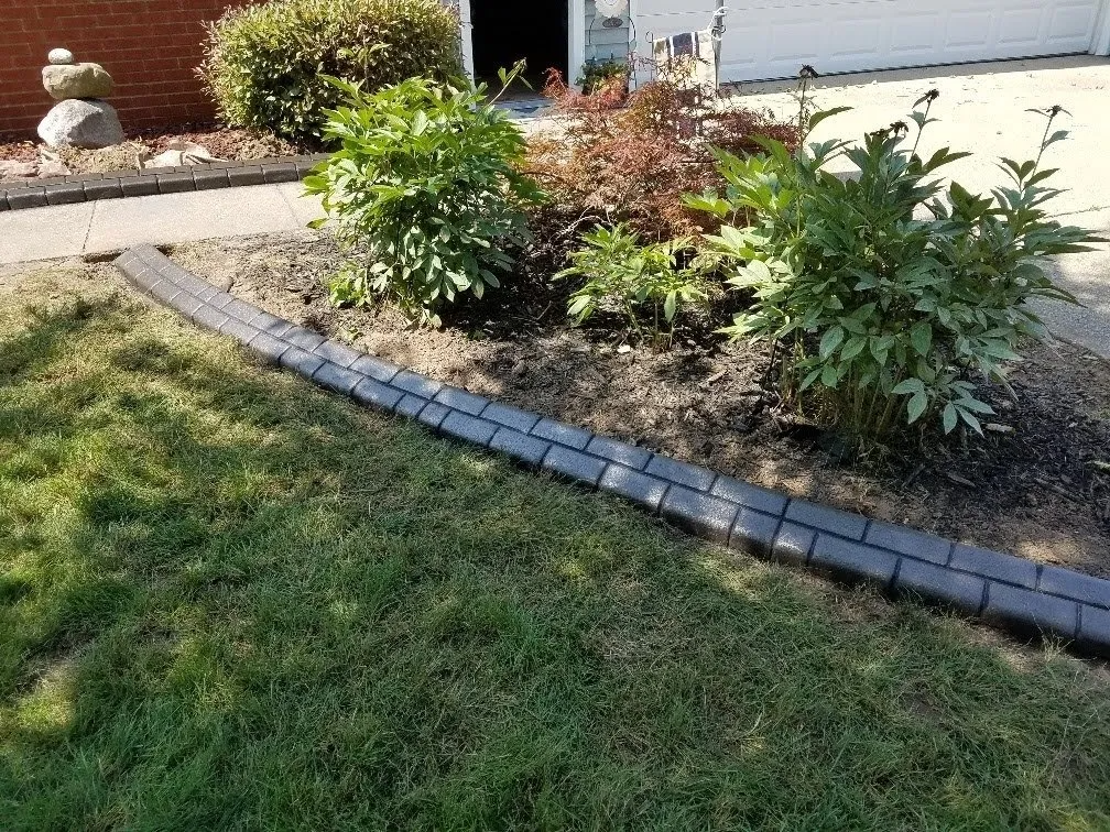 A garden bed bordered by gray stone pavers next to a green lawn and a brick building.
