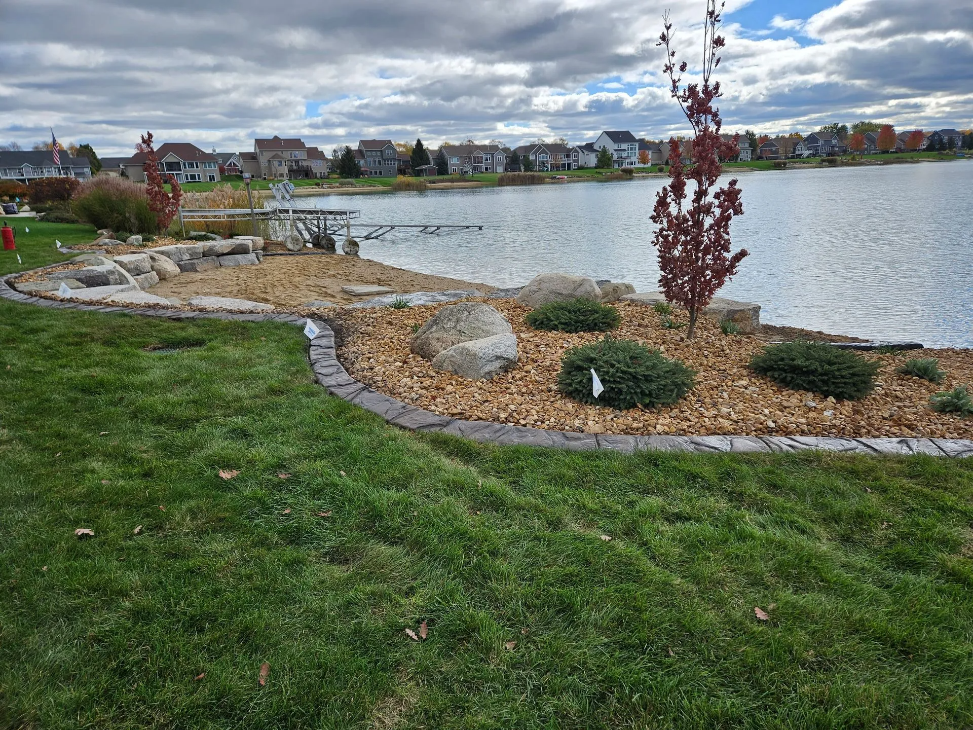 A landscaped garden bed with rocks and small shrubs, bordered by stone, overlooking a lake with houses in the distance.