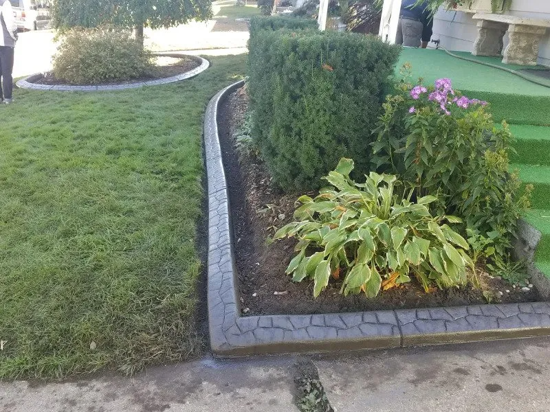 Stamped concrete landscape edging separating a green lawn and mulch bed with shrubs and flowers.