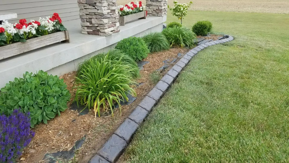 A landscaping bed with shrubs and a flower box along a stone-columned porch, bordered by gray pavers against a green lawn.