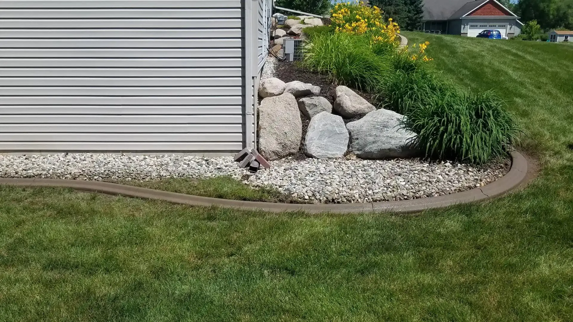 A manicured concrete curb borders a home’s side garden bed with grey siding, decorative boulders, and green plants.