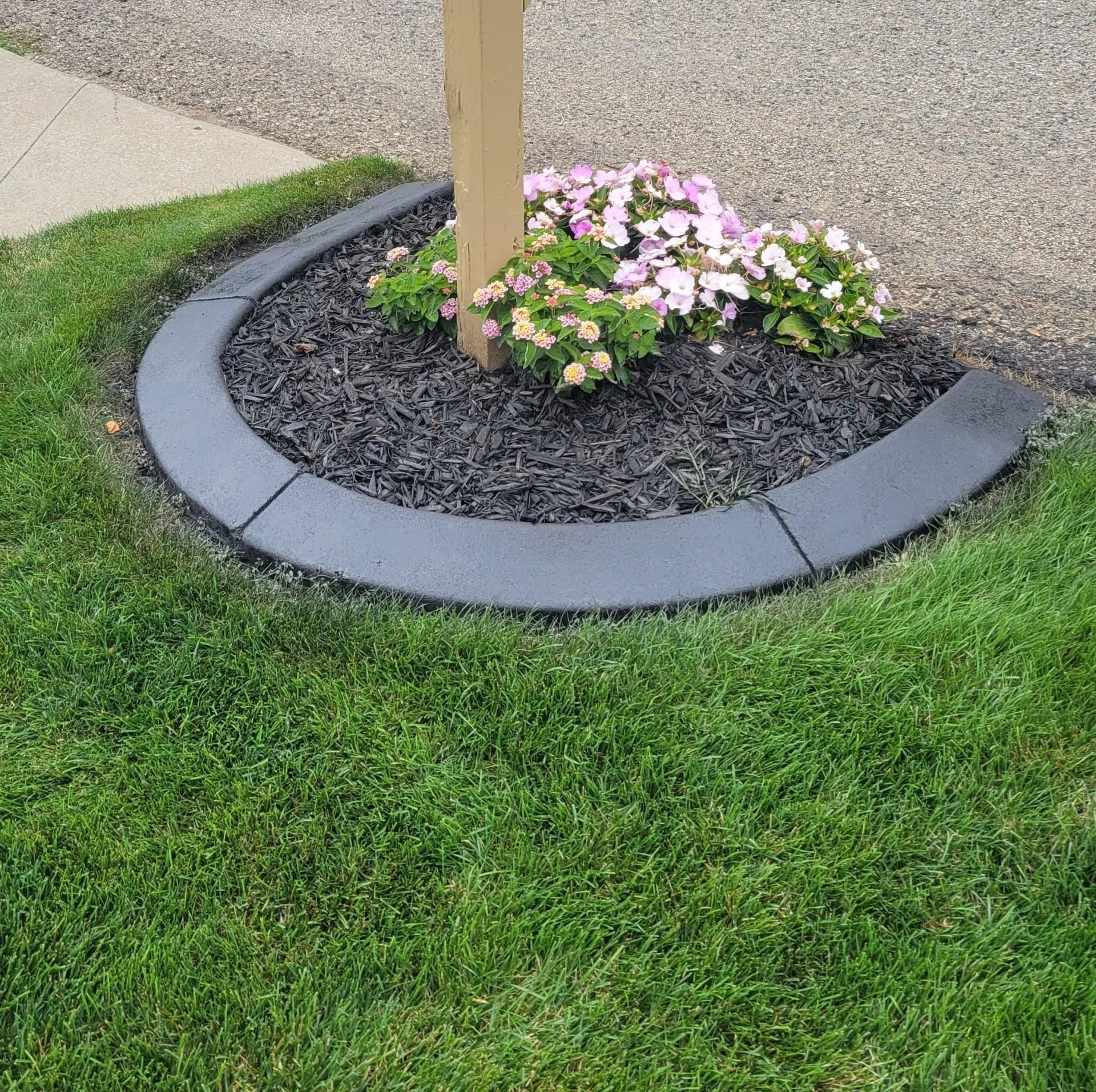 A curved, dark grey landscape border surrounds mulch and pink flowers at the base of a post in a grassy yard.