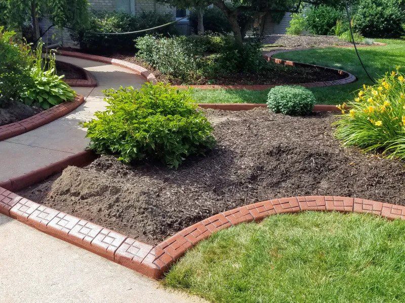 A curved garden bed with dark mulch, green plants, and patterned red brick edging bordering a walkway and lawn.