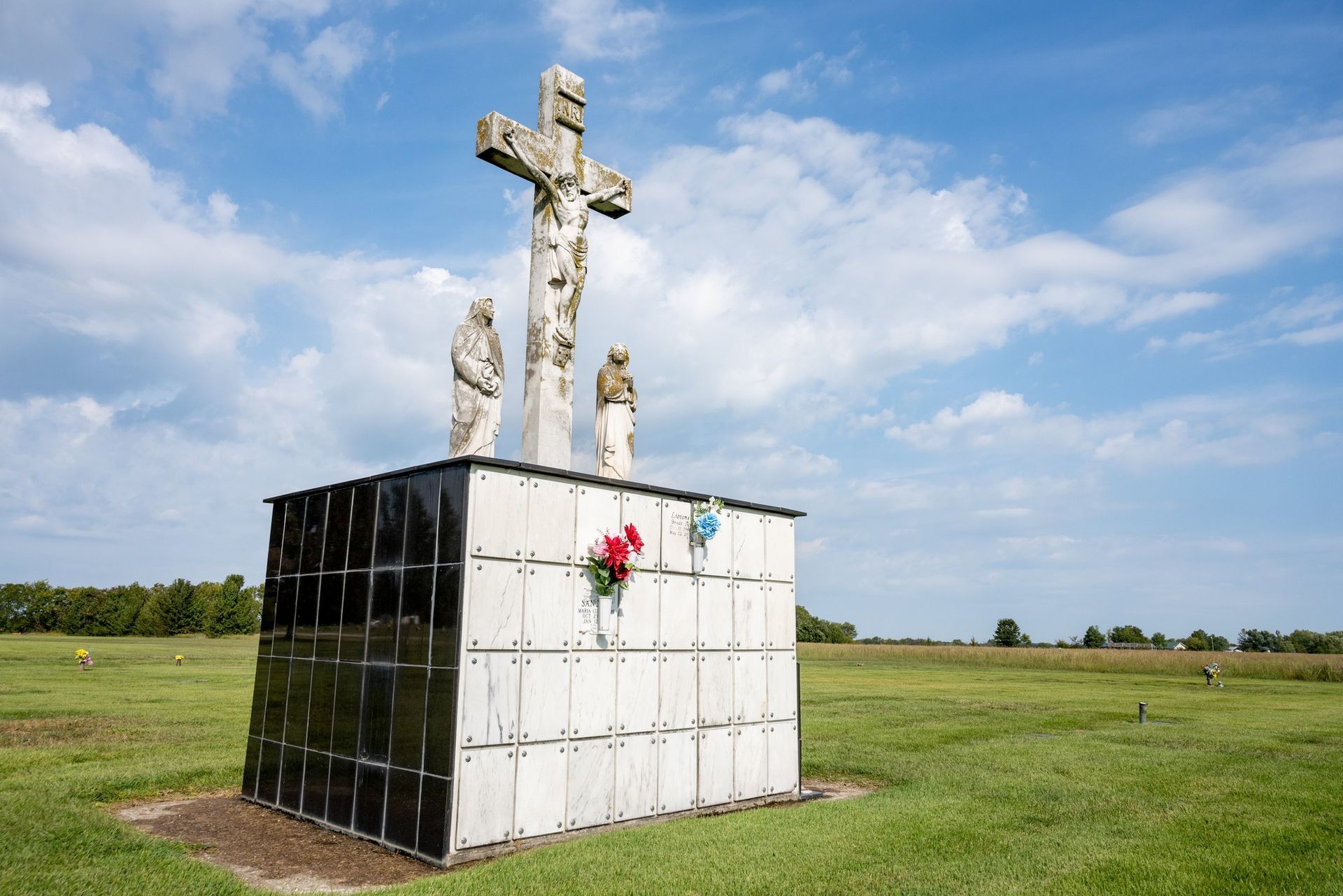 White memorial cross on a tiled pedestal in a grassy field under a blue sky