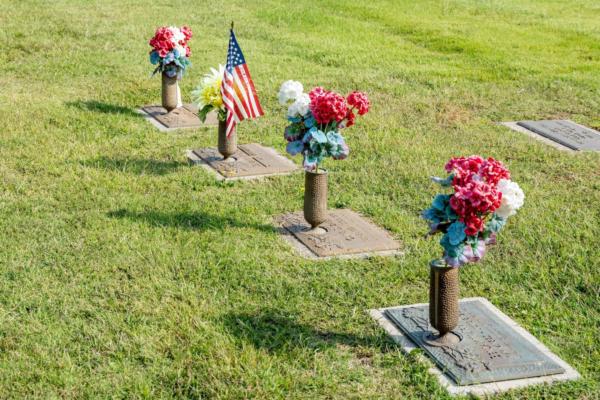 Small decorated graves with flower arrangements and an American flag in a grassy cemetery.