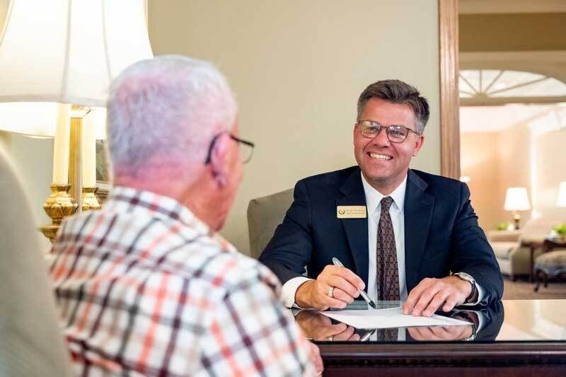 Two men in a business meeting at a table, one smiling and taking notes in a bright office.